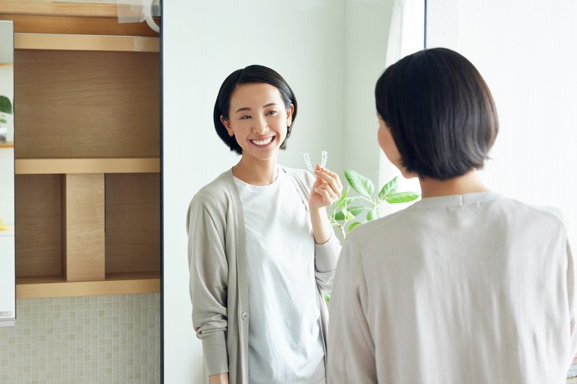 鏡の前で、透明なマウスピースを指先に持ち、笑顔を見せる女性の写真